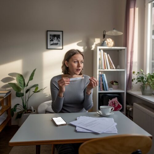 Junge Frau in einer deutschen Wohnung hält positiven Schwangerschaftstest und hat besorgten Ausdruck. Tisch mit Teetasse und Smartphone.