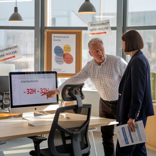 Mann mit Checkhemd zeigt auf Computeranwendung mit negativen Arbeitsstunden, Frau mit Blazer hält ungesigntes Dokument.